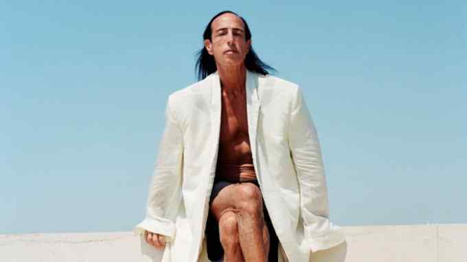 A photograph of designer Rick Owens, wearing a long white jacket but with bare chest and bare legs