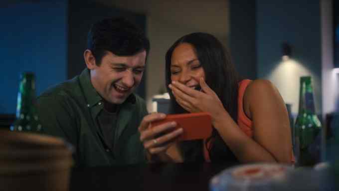A dark-haired man wearing a forest-green shirt and a brown-haired woman wearing an orange vest both laugh whilst looking at smartphone