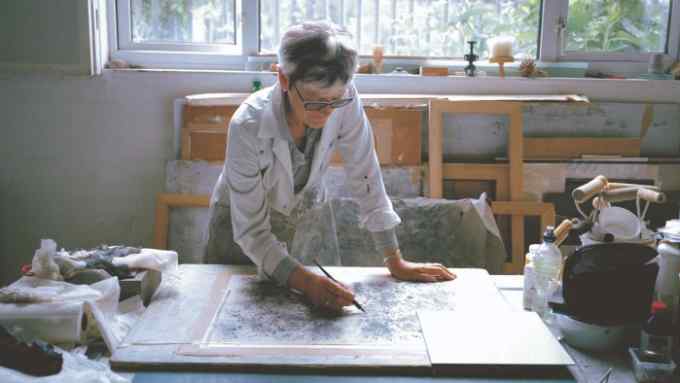 A middle-aged woman with short grey hair, dark glasses and a white shirt is portrayed painting in a naturally lit studio filled with wooden frames, textiles and paint rollers.
