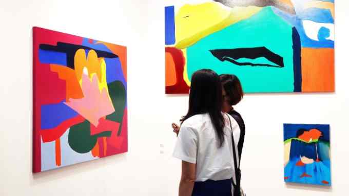 Two people look at brightly-coloured semi-abstract paintings on the white walls of an art fair
