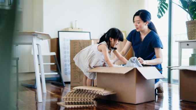 Young Asian family moving into a new apartment. Little daughter helping her mother to pack/unpack belongings into cardboard box in the living room