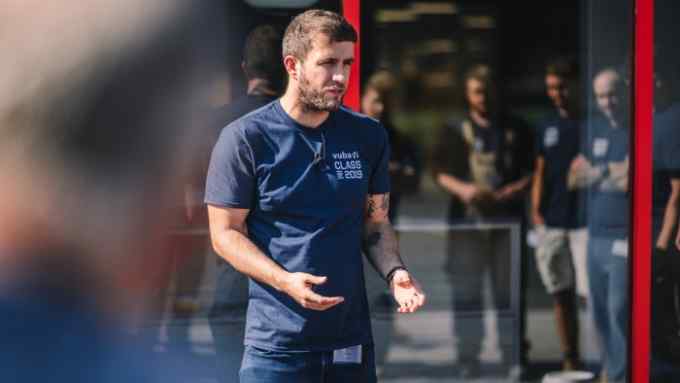 The image features a man in the foreground speaking and gesturing with his hands. He is wearing a dark blue t-shirt, has short, dark hair, a trimmed beard, and a tattoo on his left arm.