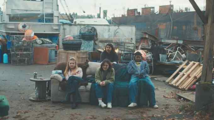 Three teenage girls sit on an old sofa in a messy junkyard. A fourth girl leans against the back of the sofa.